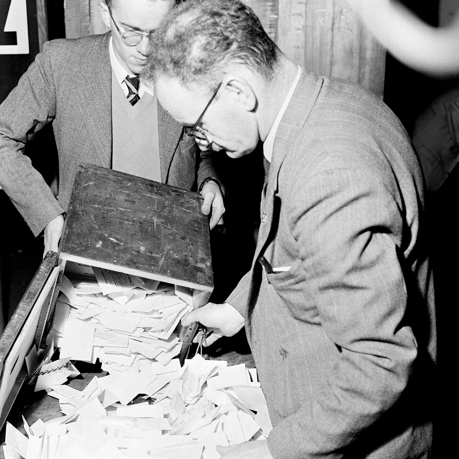 Two men empty folded votes out of a wooden ballot box for the 1954 election. Two men empty folded votes out of a wooden ballot box for the 1954 election.