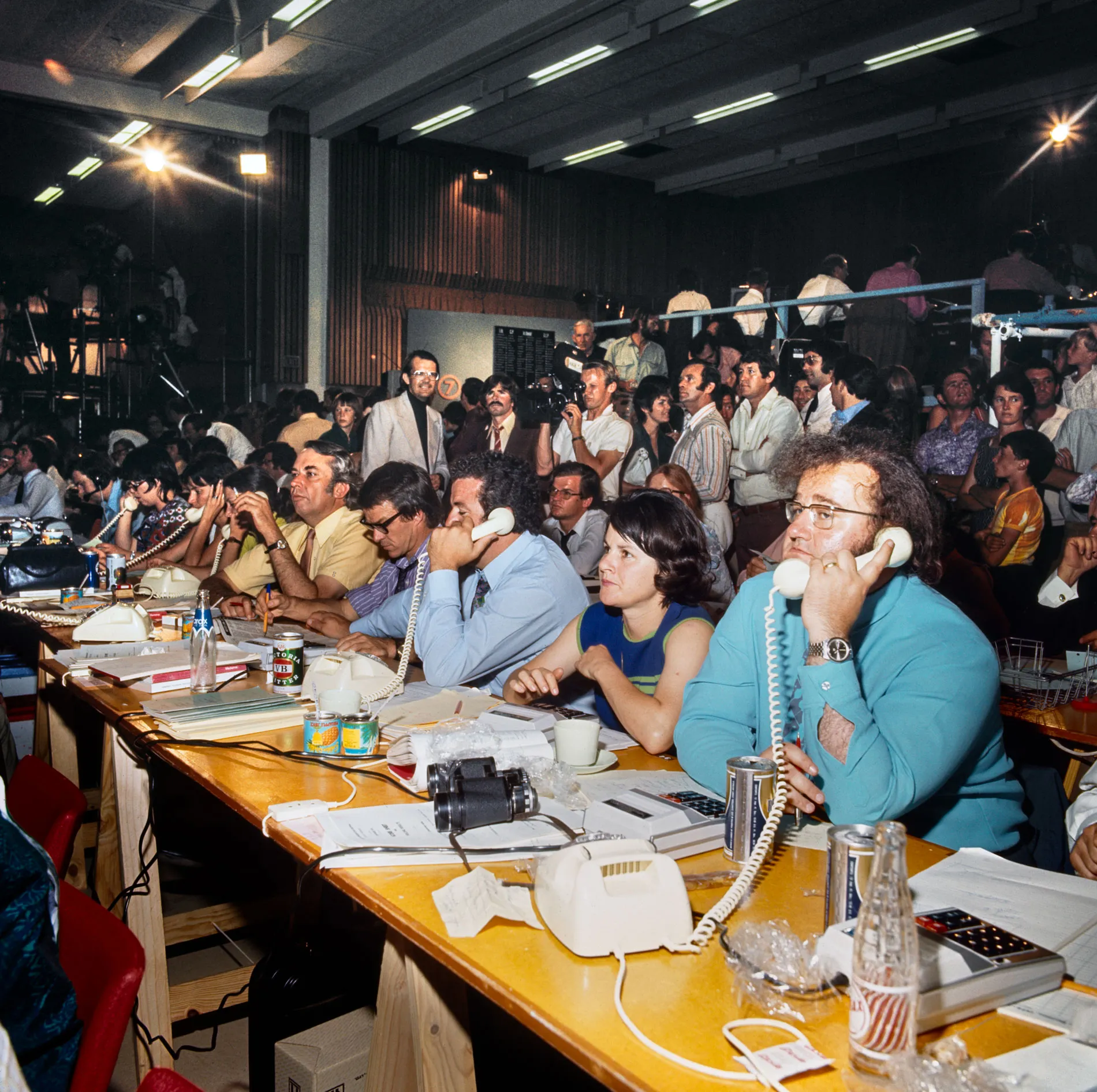 A crowd of men and women sit at tables with phones and stand holding cameras in the tally room at the 1972 election. A crowd of men and women sit at tables with phones and stand holding cameras in the tally room at the 1972 election.