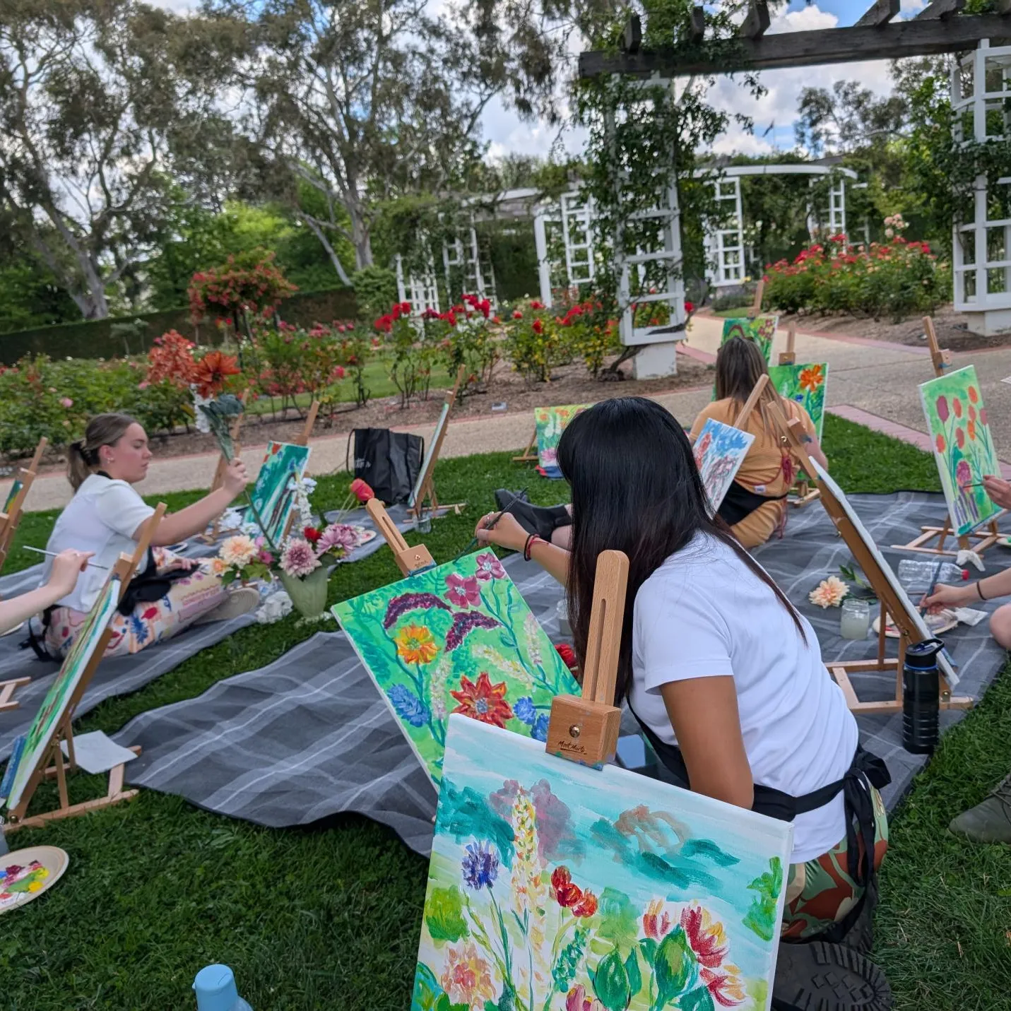 People sit on the grass in the rose gardens at Old Parliament House painting floral artworks.