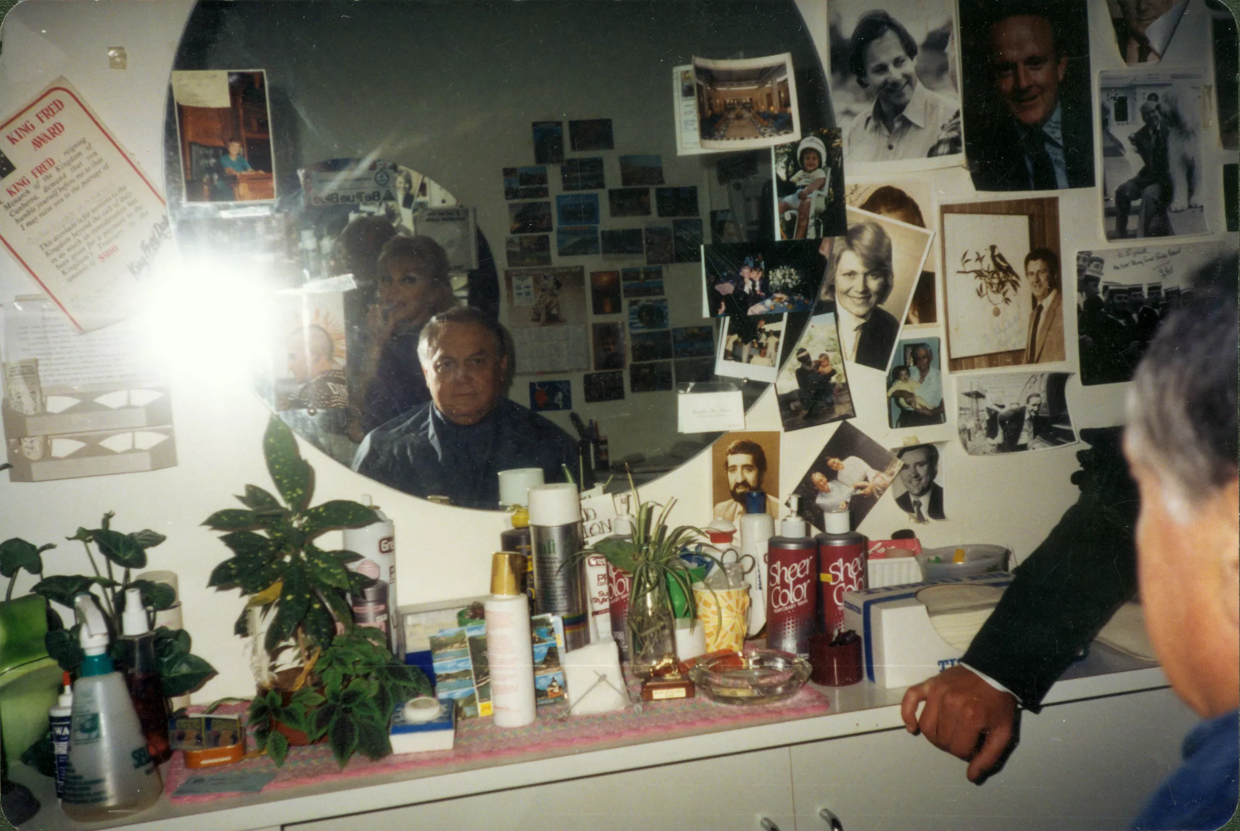 This colour photograph depicts the inside of Lizzie Scott’s salon. Gary Nehl MP sits in the barber’s chair with Lizzie standing directly behind him. They are reflected in a circular mirror which is mounted on the wall. Under the mirror, there is a counter which is littered with pot plants, hair spray, tissues, scissors, spray bottles, creams, postcards and an ashtray. Surrounding the mirror, are photographs of people in colour and black and white. This colour photograph depicts the inside of Lizzie Scott’s salon. Gary Nehl MP sits in the barber’s chair with Lizzie standing directly behind him. They are reflected in a circular mirror which is mounted on the wall. Under the mirror, there is a counter which is littered with pot plants, hair spray, tissues, scissors, spray bottles, creams, postcards and an ashtray. Surrounding the mirror, are photographs of people in colour and black and white.