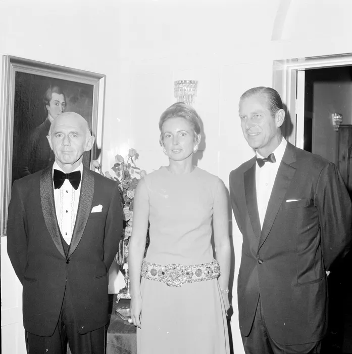 This black and white group photograph captures Prime Minister McMahon in a tuxedo with ruffled white shirt and black velvet bow tie standing next to his wife Sonia who is wearing a sleeveless light-coloured dress with heavily jewelled belt. Her long blonde hair is piled up in a chignon and she is wearing large earrings. To her left is Prince Philip in a more restrained tuxedo and bow tie. The group are standing in an elegant room and there is a flower arrangement and oil painting in the background. This black and white group photograph captures Prime Minister McMahon in a tuxedo with ruffled white shirt and black velvet bow tie standing next to his wife Sonia who is wearing a sleeveless light-coloured dress with heavily jewelled belt. Her long blonde hair is piled up in a chignon and she is wearing large earrings. To her left is Prince Philip in a more restrained tuxedo and bow tie. The group are standing in an elegant room and there is a flower arrangement and oil painting in the background.