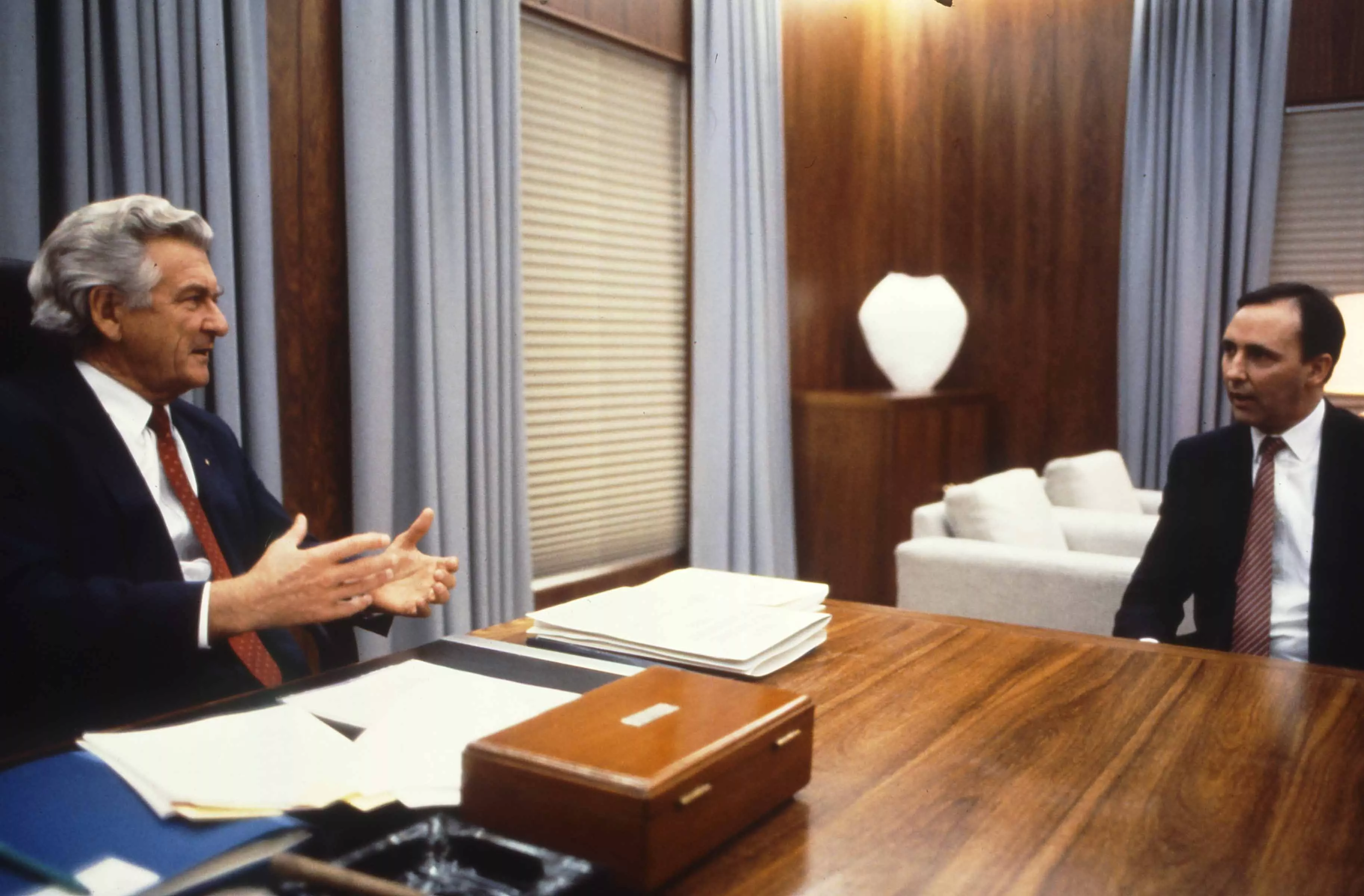 This colour photograph captures a conversation between Prime Minister Bob Hawke seated on the left and Treasurer Paul Keating seated on the right. The photo shows the corner of the desk with Hawke sitting behind the desk while Keating sits to the side with both men facing each other. Both are dressed in black suits with white shirts and red ties contrasting the pale blue curtains in the background. This colour photograph captures a conversation between Prime Minister Bob Hawke seated on the left and Treasurer Paul Keating seated on the right. The photo shows the corner of the desk with Hawke sitting behind the desk while Keating sits to the side with both men facing each other. Both are dressed in black suits with white shirts and red ties contrasting the pale blue curtains in the background.