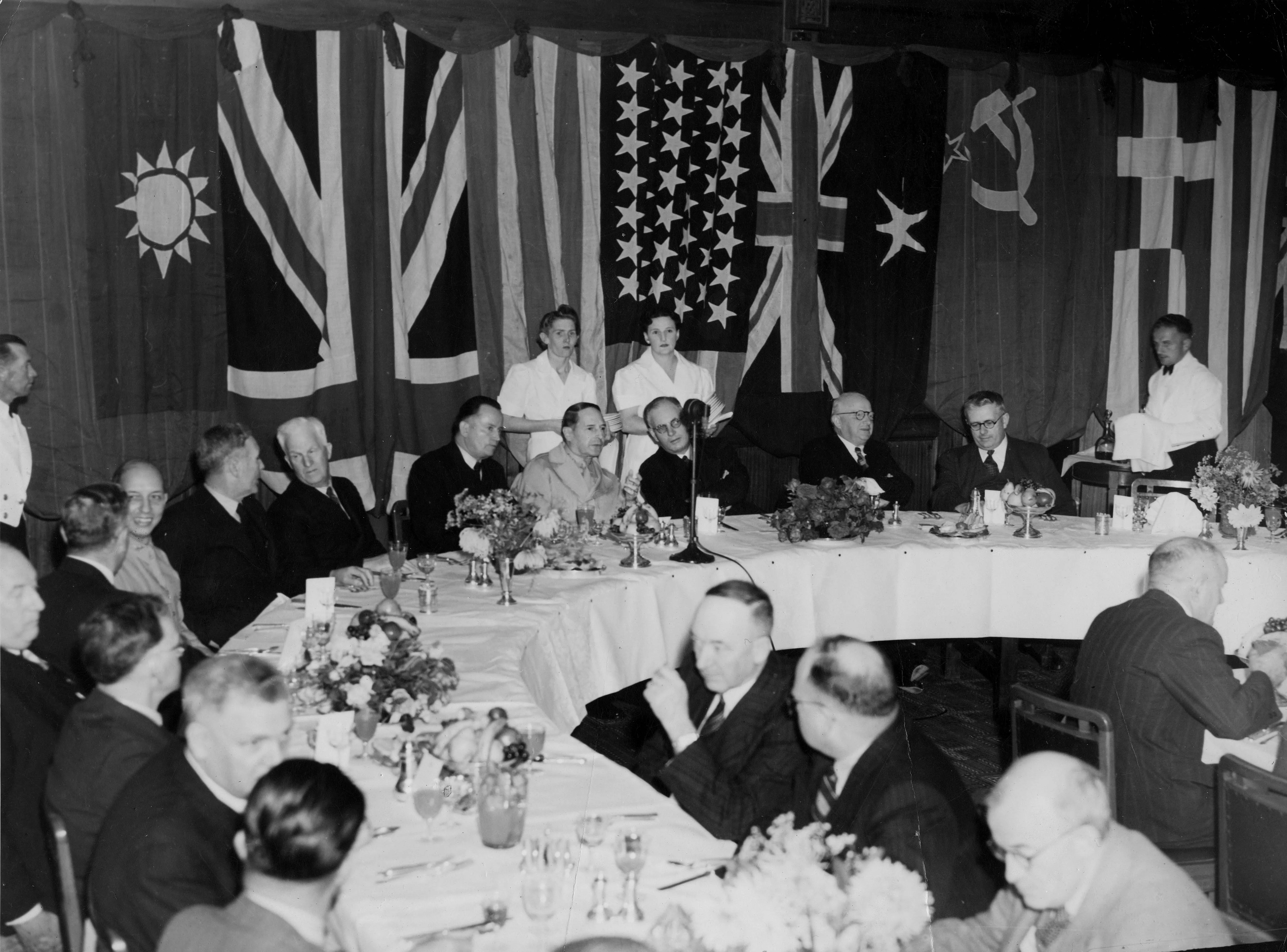 This black and white photograph shows a section of a dining table in the Members’ Dining Room with national flags hanging in the background. General Douglas MacArthur is seated at the head of the table with a microphone in front of him; he is between Deputy Leader of the ALP Frank Forde and Prime Minister John Curtin. Other politicians are seated at the table which is set with white linen, silverware, glassware, crockery and flower arrangements. Uniformed waiters and waitresses stand behind the diners. This black and white photograph shows a section of a dining table in the Members’ Dining Room with national flags hanging in the background. General Douglas MacArthur is seated at the head of the table with a microphone in front of him; he is between Deputy Leader of the ALP Frank Forde and Prime Minister John Curtin. Other politicians are seated at the table which is set with white linen, silverware, glassware, crockery and flower arrangements. Uniformed waiters and waitresses stand behind the diners.