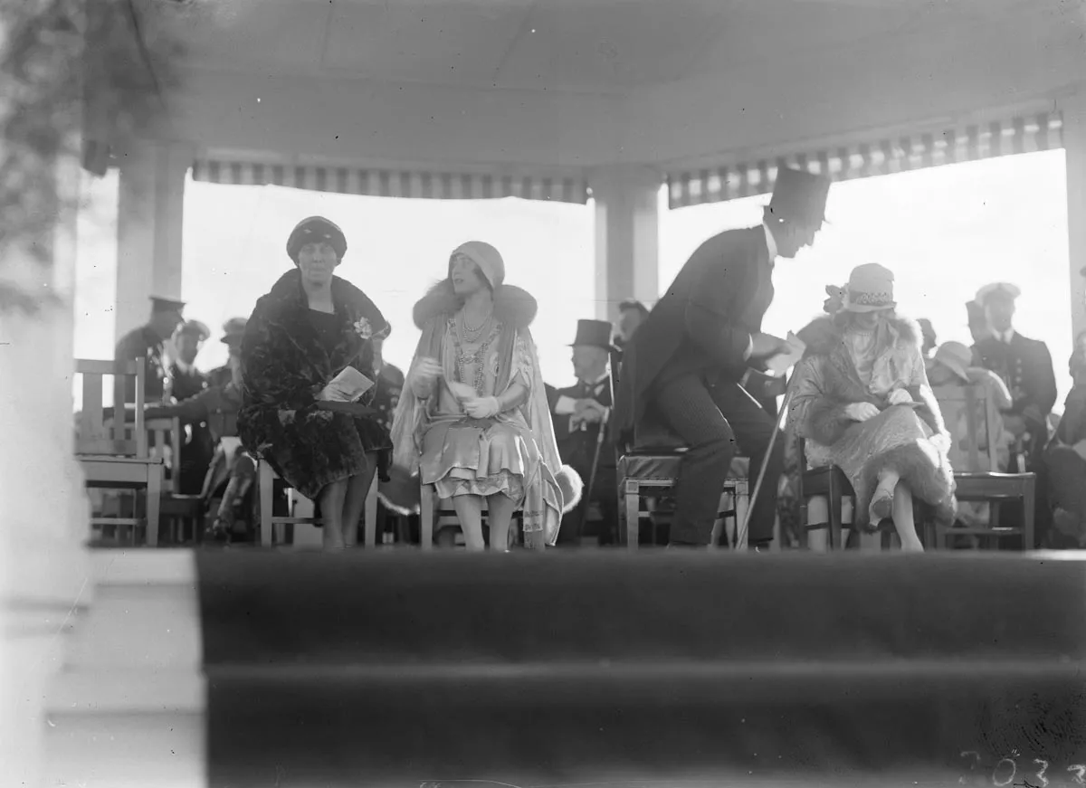 This black and white photograph lets us peek over the edge of the carpeted floor of the white timber Royal dais constructed at York Park for the official party to watch the Troop Review. On the left, a seated Mrs Ethel Bruce chats with the Duchess of York. Ethel is dressed in a dark, luxurious fur coat with cloche hat and gloves. The Duchess is richly dressed in a pale-coloured lustrous dress with elaborate embroidery and a fur-trimmed cape, cloche hat and white gloves. Prime Minister Bruce, in top hat and This black and white photograph lets us peek over the edge of the carpeted floor of the white timber Royal dais constructed at York Park for the official party to watch the Troop Review. On the left, a seated Mrs Ethel Bruce chats with the Duchess of York. Ethel is dressed in a dark, luxurious fur coat with cloche hat and gloves. The Duchess is richly dressed in a pale-coloured lustrous dress with elaborate embroidery and a fur-trimmed cape, cloche hat and white gloves. Prime Minister Bruce, in top hat and