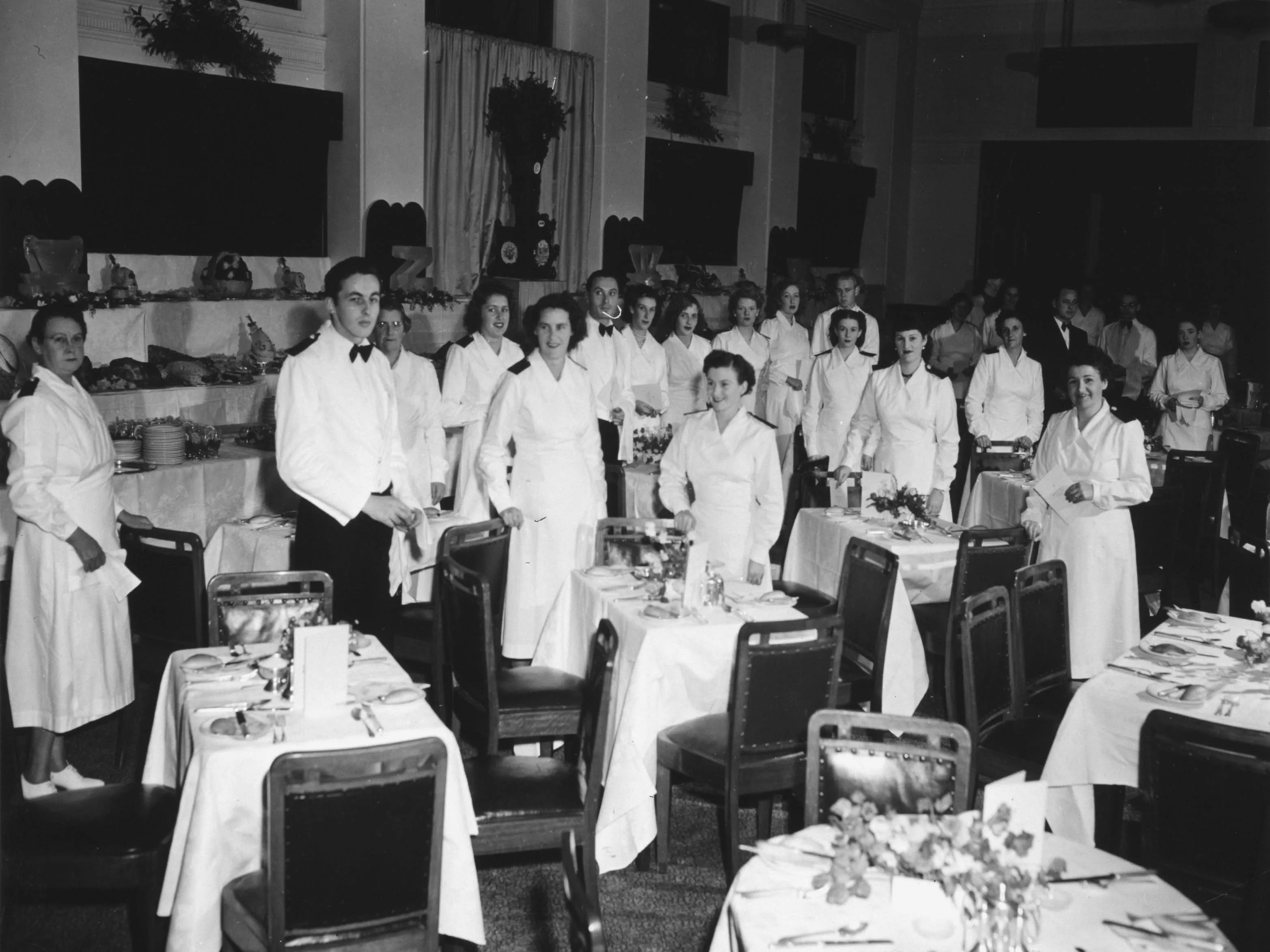This black and white photograph captures the moment before a dinner celebrating the 50th anniversary of Federation in the Members’ Dining Room at Parliament House. Small dining tables seating two to four guests are set up in the foreground with white linen tablecloths and napkins, silverware, crockery, bread rolls, floral arrangements and menus. In the background are tiered tables laden with food and crockery and topped with ice sculptures, centrepieces and floral arrangements. In the midground, 23 members This black and white photograph captures the moment before a dinner celebrating the 50th anniversary of Federation in the Members’ Dining Room at Parliament House. Small dining tables seating two to four guests are set up in the foreground with white linen tablecloths and napkins, silverware, crockery, bread rolls, floral arrangements and menus. In the background are tiered tables laden with food and crockery and topped with ice sculptures, centrepieces and floral arrangements. In the midground, 23 members