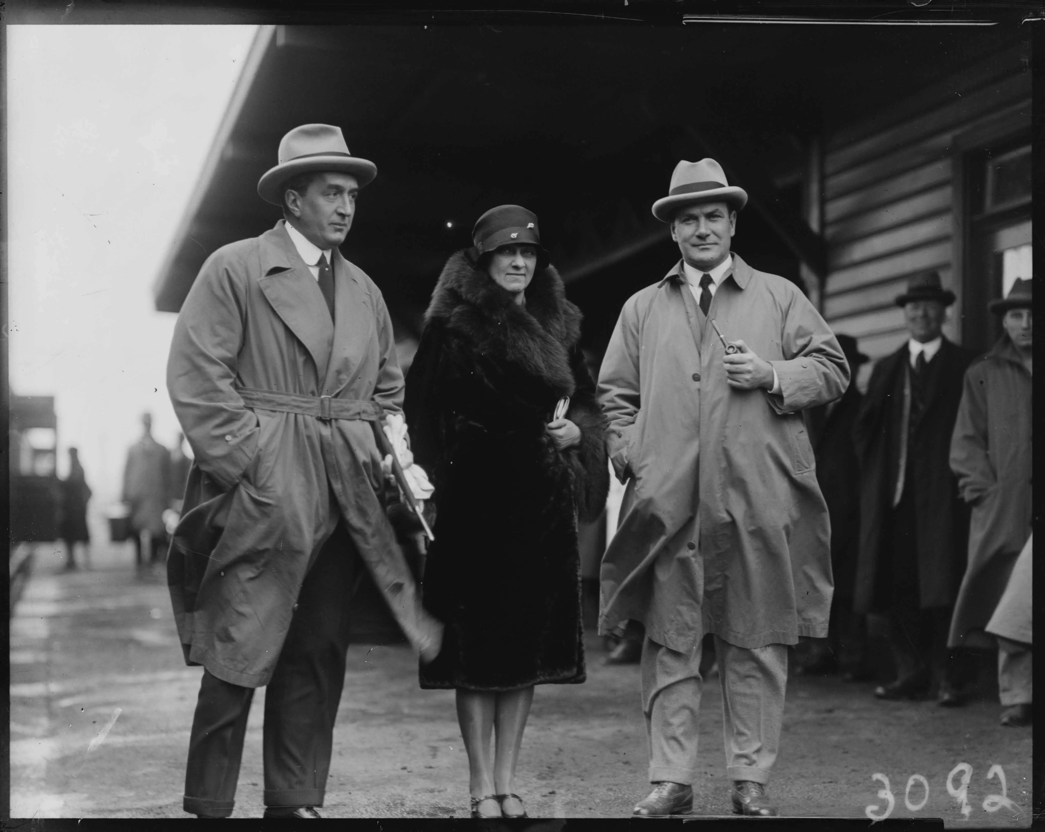 This black and white photograph shows three individuals standing on the platform at the weatherboard Canberra Railway Station on 8 May 1927. In the distance you can see the open doors of a train and people walking down the platform with luggage. Some other people are sheltering close to the railway station building. The weather looks cool and windy. On the left is Prime Minister Stanley Melbourne Bruce dressed in a dark suit with white shirt and dark tie over which is a belted trench coat in a lighter shade This black and white photograph shows three individuals standing on the platform at the weatherboard Canberra Railway Station on 8 May 1927. In the distance you can see the open doors of a train and people walking down the platform with luggage. Some other people are sheltering close to the railway station building. The weather looks cool and windy. On the left is Prime Minister Stanley Melbourne Bruce dressed in a dark suit with white shirt and dark tie over which is a belted trench coat in a lighter shade