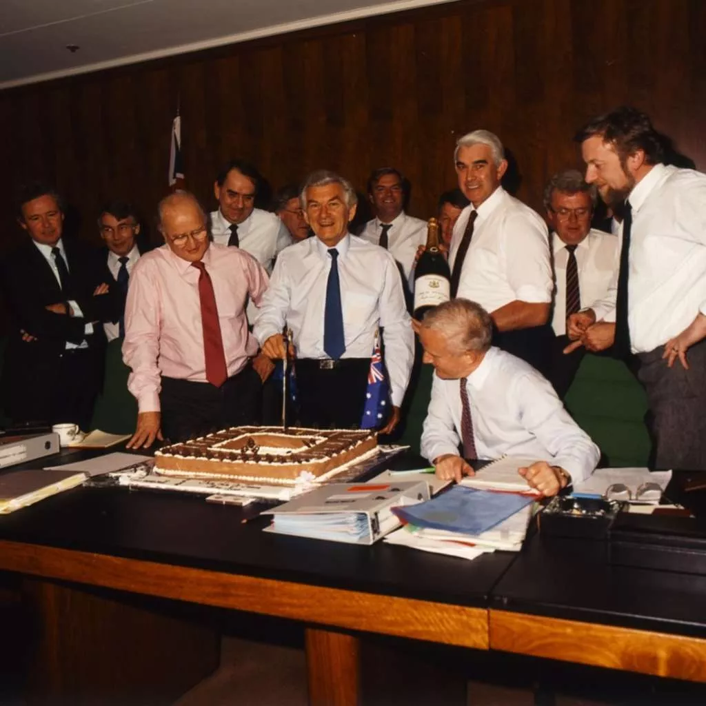 Prime Minister Bob Hawke stands and cuts a square cake, surrounded by a group of men all smiling. Prime Minister Bob Hawke stands and cuts a square cake, surrounded by a group of men all smiling.