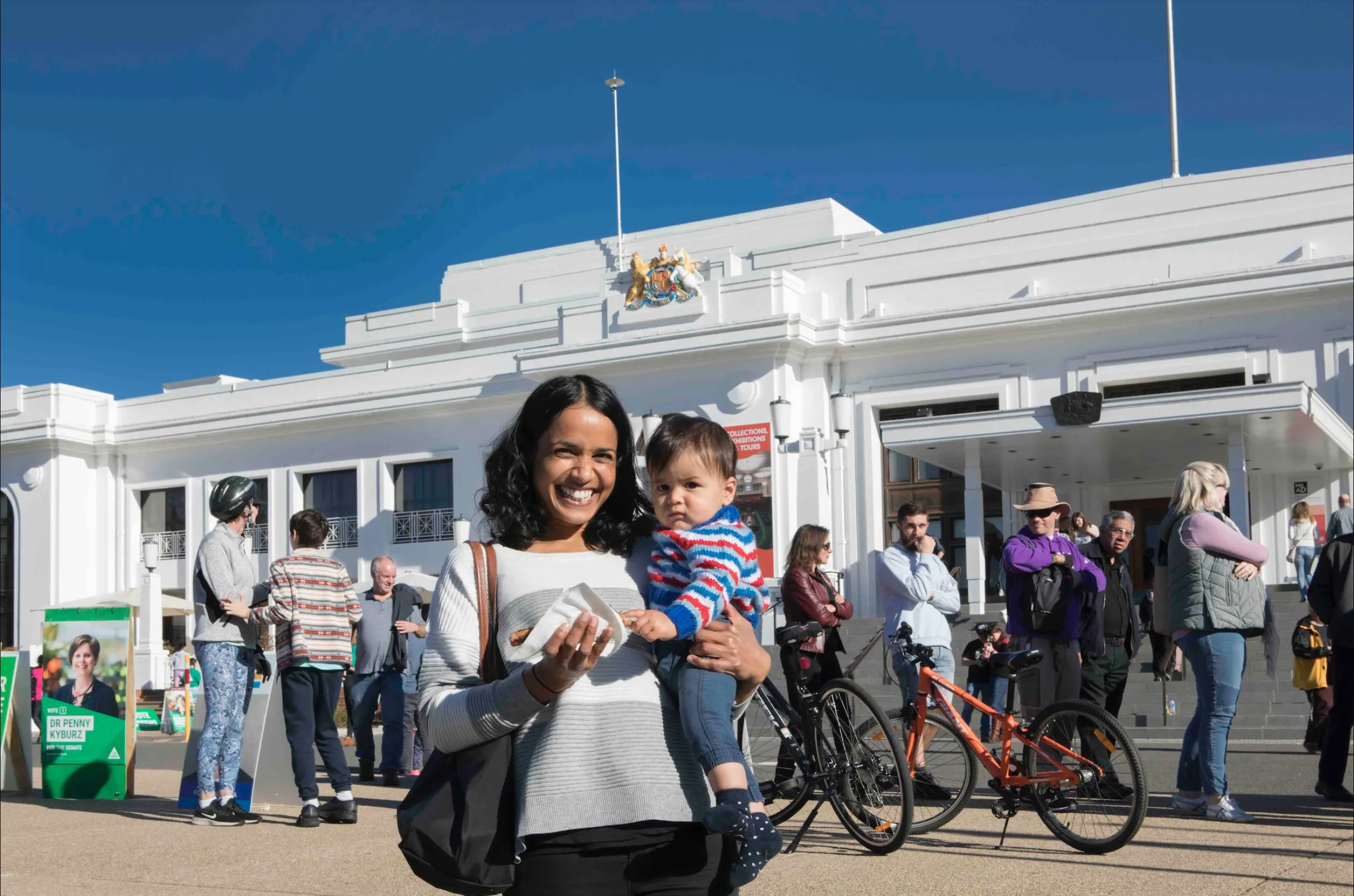 A mother holds her baby and smiles at the camera at the front of Old Parliament House. A row of people waiting to vote are behind her. She holds a sausage sandwich. A mother holds her baby and smiles at the camera at the front of Old Parliament House. A row of people waiting to vote are behind her. She holds a sausage sandwich.