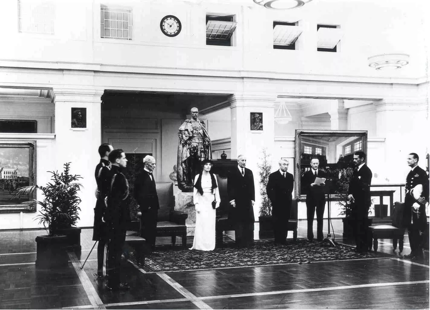 This black and white photograph shows a group of people standing at attention in King’s Hall while the announcement is read. The statue of George the Fifth looms over the group. Ther are seats behind the attendees, including the Vice Regal Chair which would have been moved from the Senate Chamber. The men wear dark suits or military uniforms while the Governor-General’s wife, Lady Gowrie, wears a white formal dress, hat and gloves. This black and white photograph shows a group of people standing at attention in King’s Hall while the announcement is read. The statue of George the Fifth looms over the group. Ther are seats behind the attendees, including the Vice Regal Chair which would have been moved from the Senate Chamber. The men wear dark suits or military uniforms while the Governor-General’s wife, Lady Gowrie, wears a white formal dress, hat and gloves.