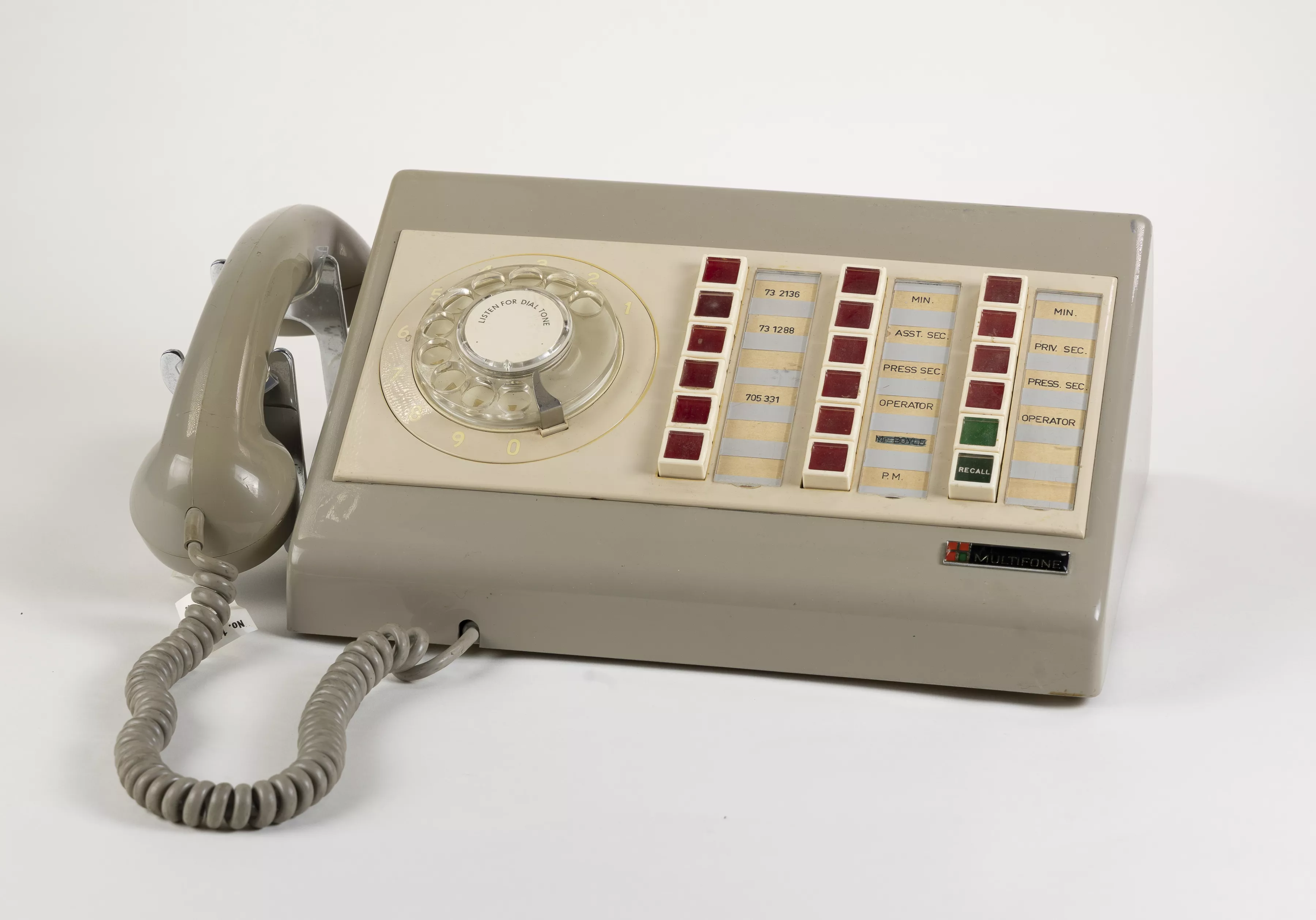 A light taupe plastic Multifone phone with 16 red buttons, 2 green buttons and a manual hand dial. The receiver sits on the side with a curly rubber cord. A light taupe plastic Multifone phone with 16 red buttons, 2 green buttons and a manual hand dial. The receiver sits on the side with a curly rubber cord.