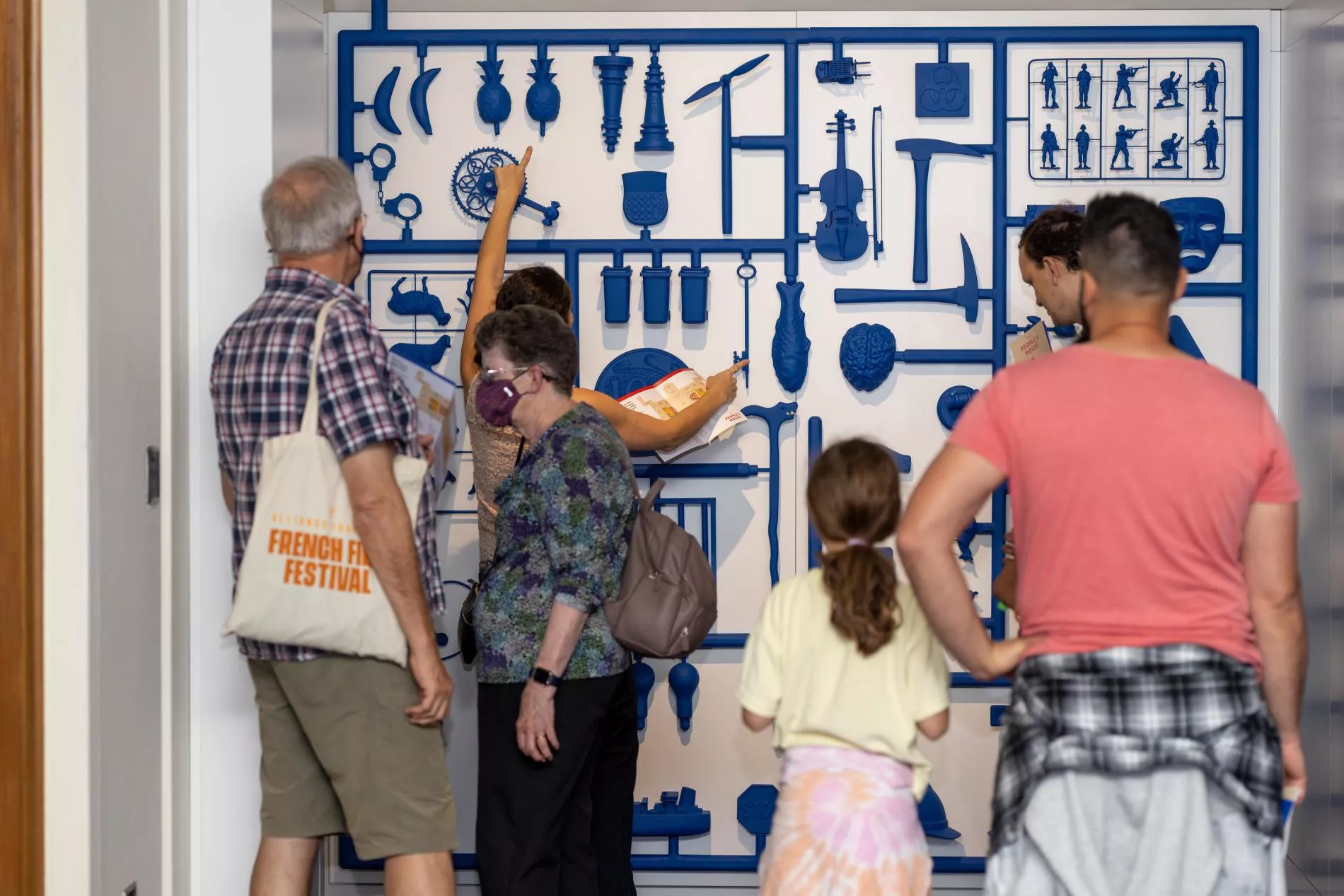 A group of visitors gather in front of a wall with a large blue installation that looks like the plastic pieces of a board game. A group of visitors gather in front of a wall with a large blue installation that looks like the plastic pieces of a board game.