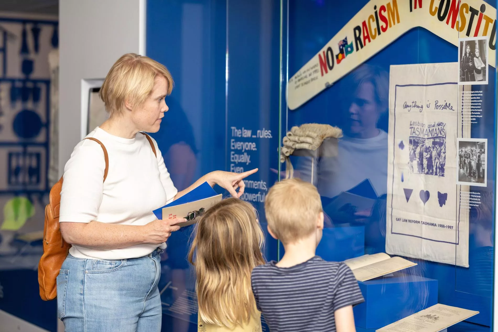 A woman standing with a boy and a girl points to a glass case with a boomerang with the words no racism in the constitution on it. A woman standing with a boy and a girl points to a glass case with a boomerang with the words no racism in the constitution on it.
