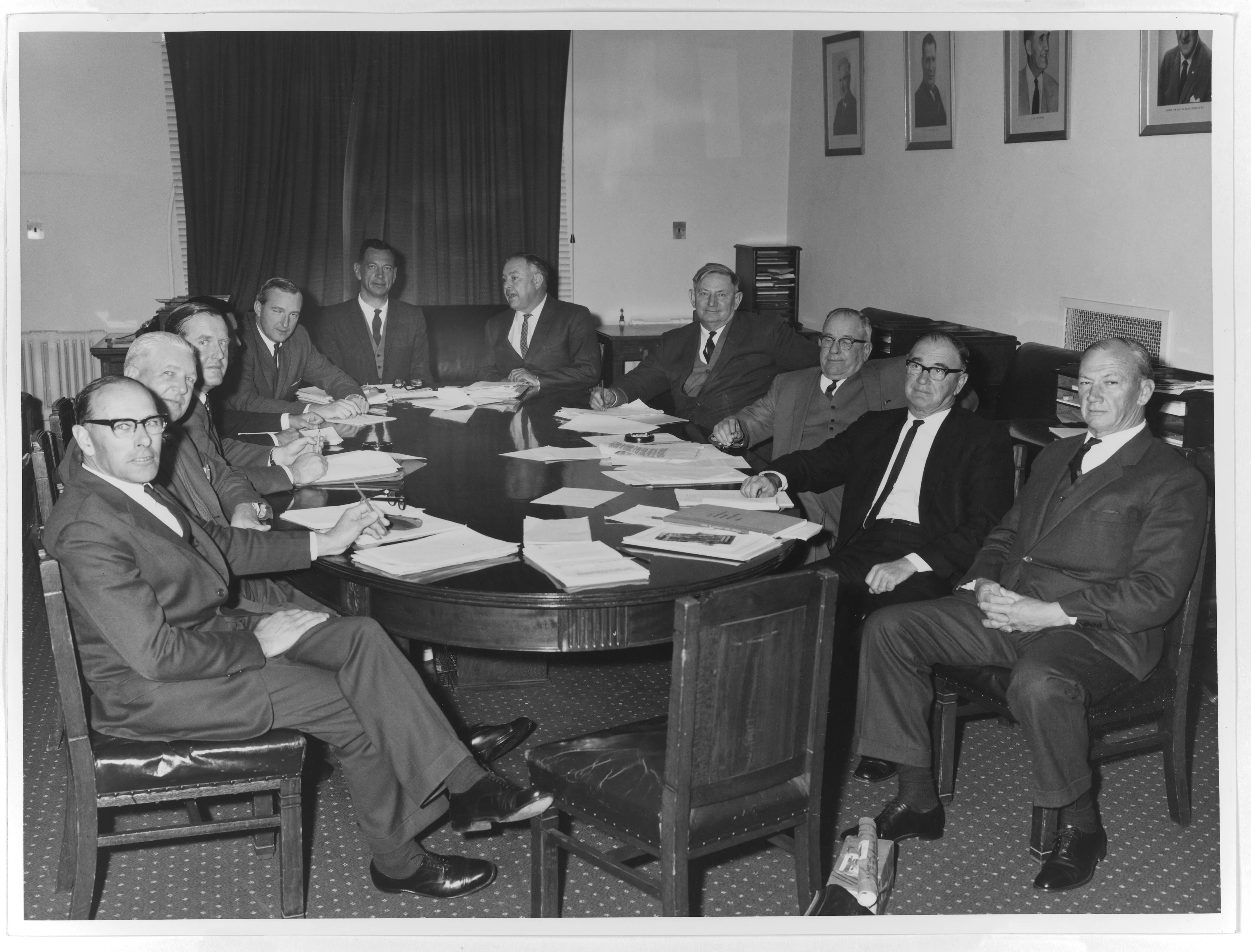 This black and white photograph shows 11 members of the Country Party Standing Committee on Drought meeting in the party room. They are seated at a large oval table covered with papers and almost all of the men have looked up to acknowledge the camera. They are uniformly dressed in dark suits, white shirts and ties and some are wearing waistcoats. In the background is a large curtained window, small desks with stationery holders and framed portraits of Country Party leaders. This black and white photograph shows 11 members of the Country Party Standing Committee on Drought meeting in the party room. They are seated at a large oval table covered with papers and almost all of the men have looked up to acknowledge the camera. They are uniformly dressed in dark suits, white shirts and ties and some are wearing waistcoats. In the background is a large curtained window, small desks with stationery holders and framed portraits of Country Party leaders.