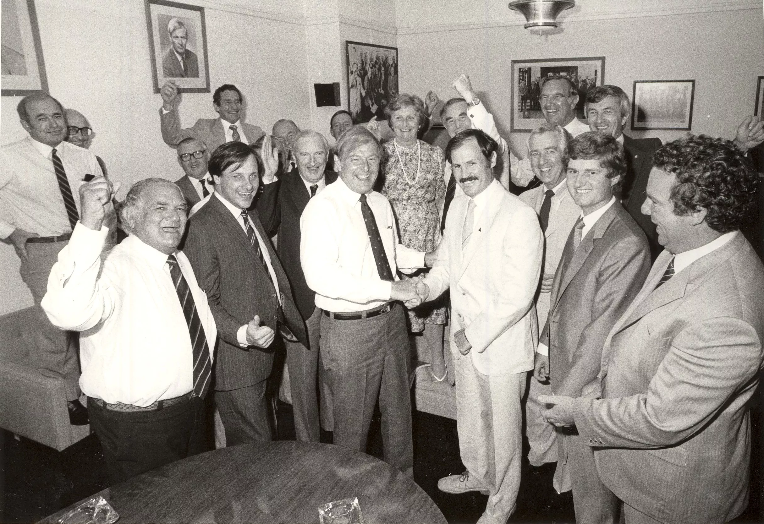 This black and white photograph captures the jubilation in the National Party Room on the first day back after the March 1983 federal election. Leader Doug Anthony shakes the hand of Noel Hicks, member for Riverina. Some of Noel’s colleagues have their hands raised in fist pumps and Flo Bjelke-Petersen is standing on a sofa with a big grin on her face. In the background are portraits of leaders and formal and informal photographs of the party. This black and white photograph captures the jubilation in the National Party Room on the first day back after the March 1983 federal election. Leader Doug Anthony shakes the hand of Noel Hicks, member for Riverina. Some of Noel’s colleagues have their hands raised in fist pumps and Flo Bjelke-Petersen is standing on a sofa with a big grin on her face. In the background are portraits of leaders and formal and informal photographs of the party.