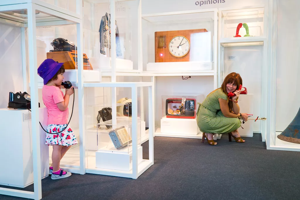 A child speaks on a phone talking to their mother surrounded by object cases in a museum with a clock, a radio and a TV. A child speaks on a phone talking to their mother surrounded by object cases in a museum with a clock, a radio and a TV.