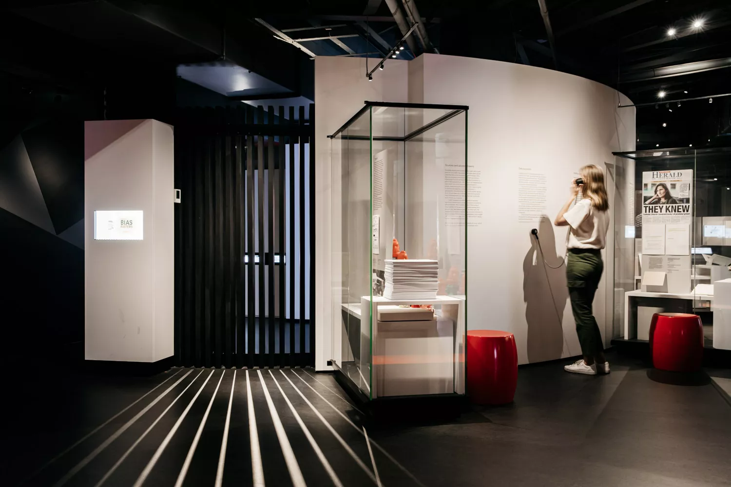 A woman listens to an audio device in an exhibition surrounded by glass panels with papers in them. A woman listens to an audio device in an exhibition surrounded by glass panels with papers in them.