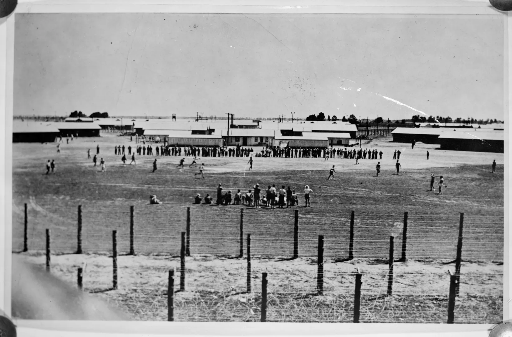 Black and white photograph of Prisoners playing soccer in an internment camp, with military buildings behind them and razor wire in the foreground. Black and white photograph of Prisoners playing soccer in an internment camp, with military buildings behind them and razor wire in the foreground.