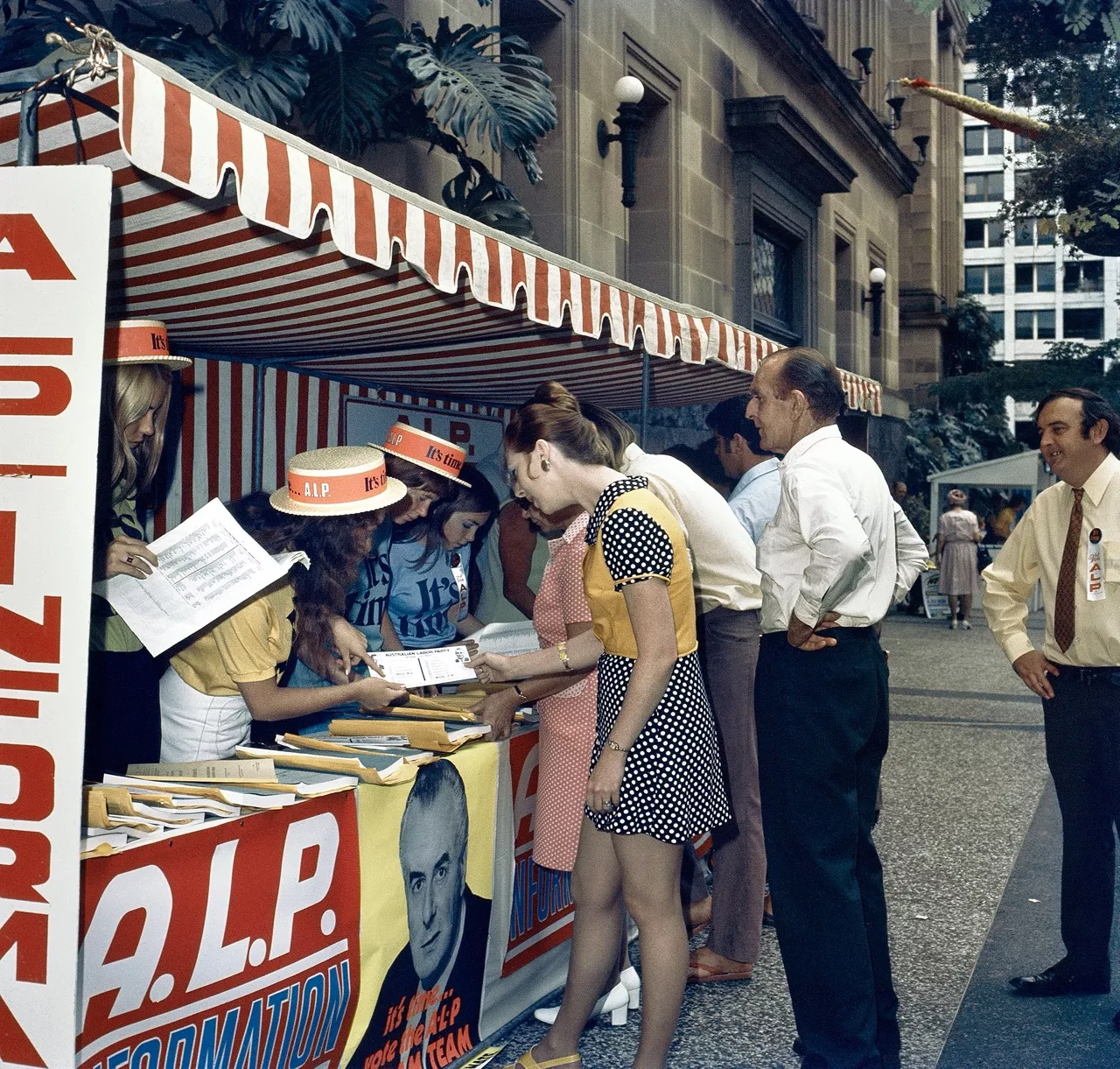 Campaign workers wearing orange bands around their hats at a booth covered in Labor bunting and poster, speaking to voters and showing them leaflets in a city street. Campaign workers wearing orange bands around their hats at a booth covered in Labor bunting and poster, speaking to voters and showing them leaflets in a city street.