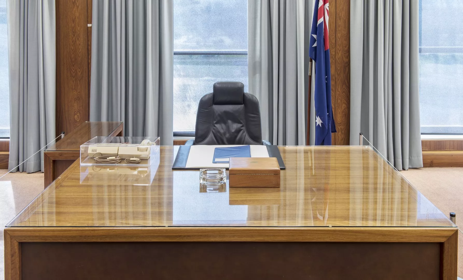 A front view of the Prime Minister's desk with a chair behind it. On the desk there is a 1980s telephone, blotter, glass ashtray and timber box. A front view of the Prime Minister's desk with a chair behind it. On the desk there is a 1980s telephone, blotter, glass ashtray and timber box.