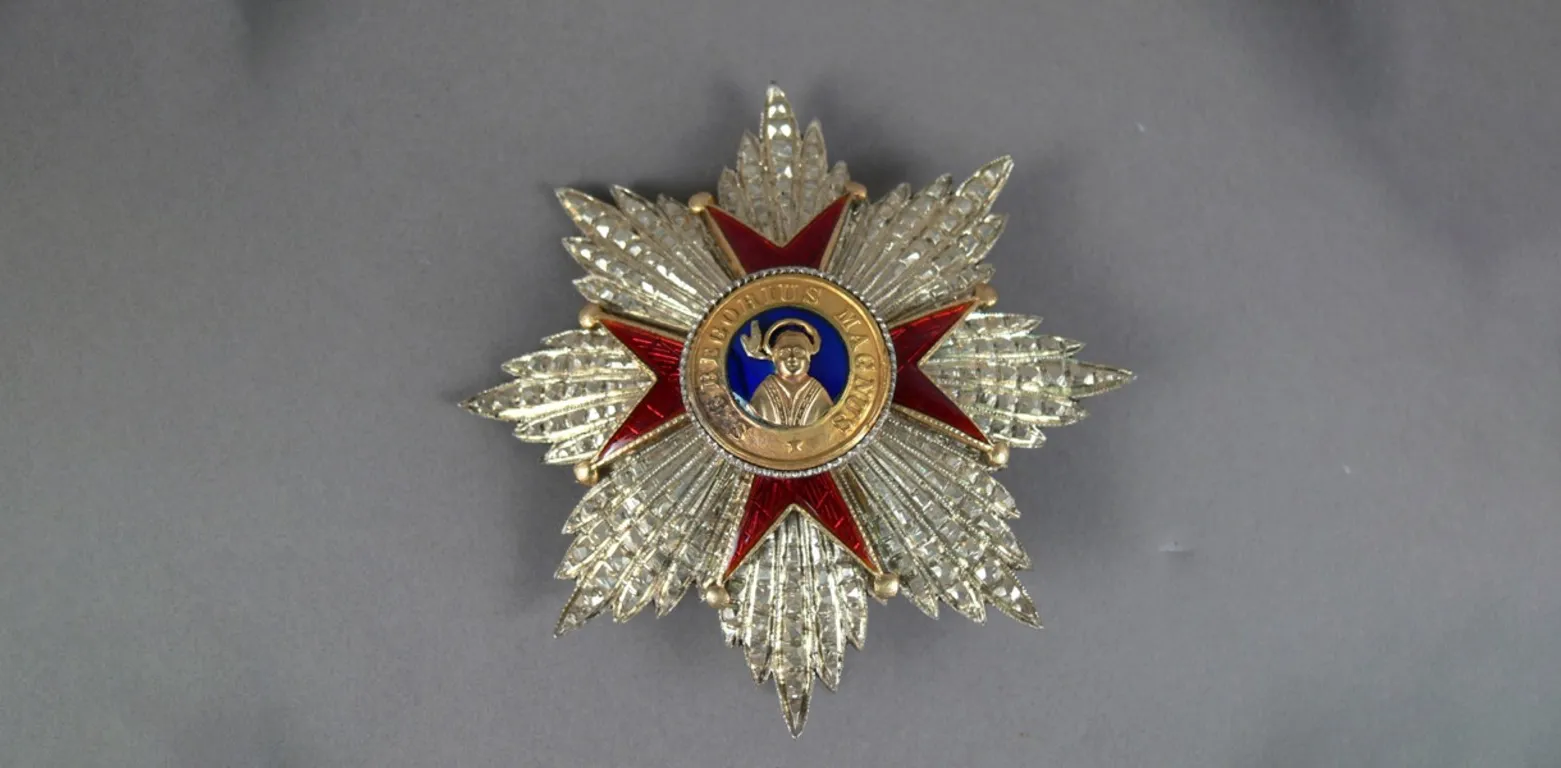 Close-up of a medal in the shape of an ornate silver star, a red cross with a gold centre and the words S. Gregorius Magnus and a golden figure. Close-up of a medal in the shape of an ornate silver star, a red cross with a gold centre and the words S. Gregorius Magnus and a golden figure.