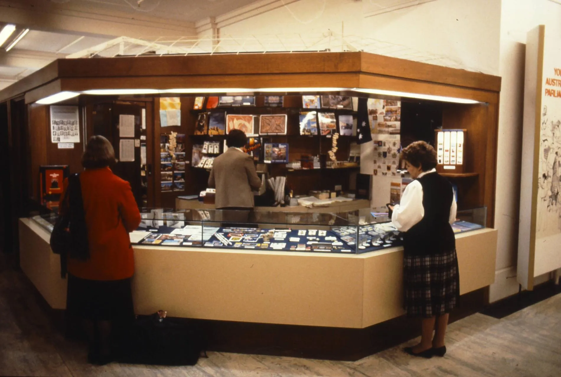 Two women stand at the glass display counter of the Old Parliament House gift shop. A man behind the counter is on a till. Two women stand at the glass display counter of the Old Parliament House gift shop. A man behind the counter is on a till.
