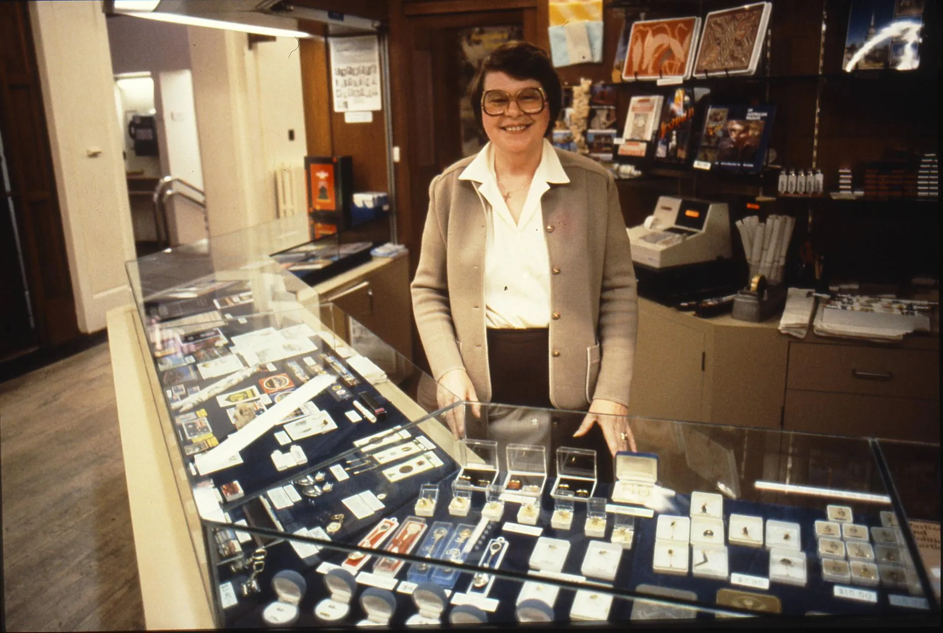 A smiling woman stands behind a glass display cabinet full of souvenirs in the gift shop at Old Parliament House. A smiling woman stands behind a glass display cabinet full of souvenirs in the gift shop at Old Parliament House.