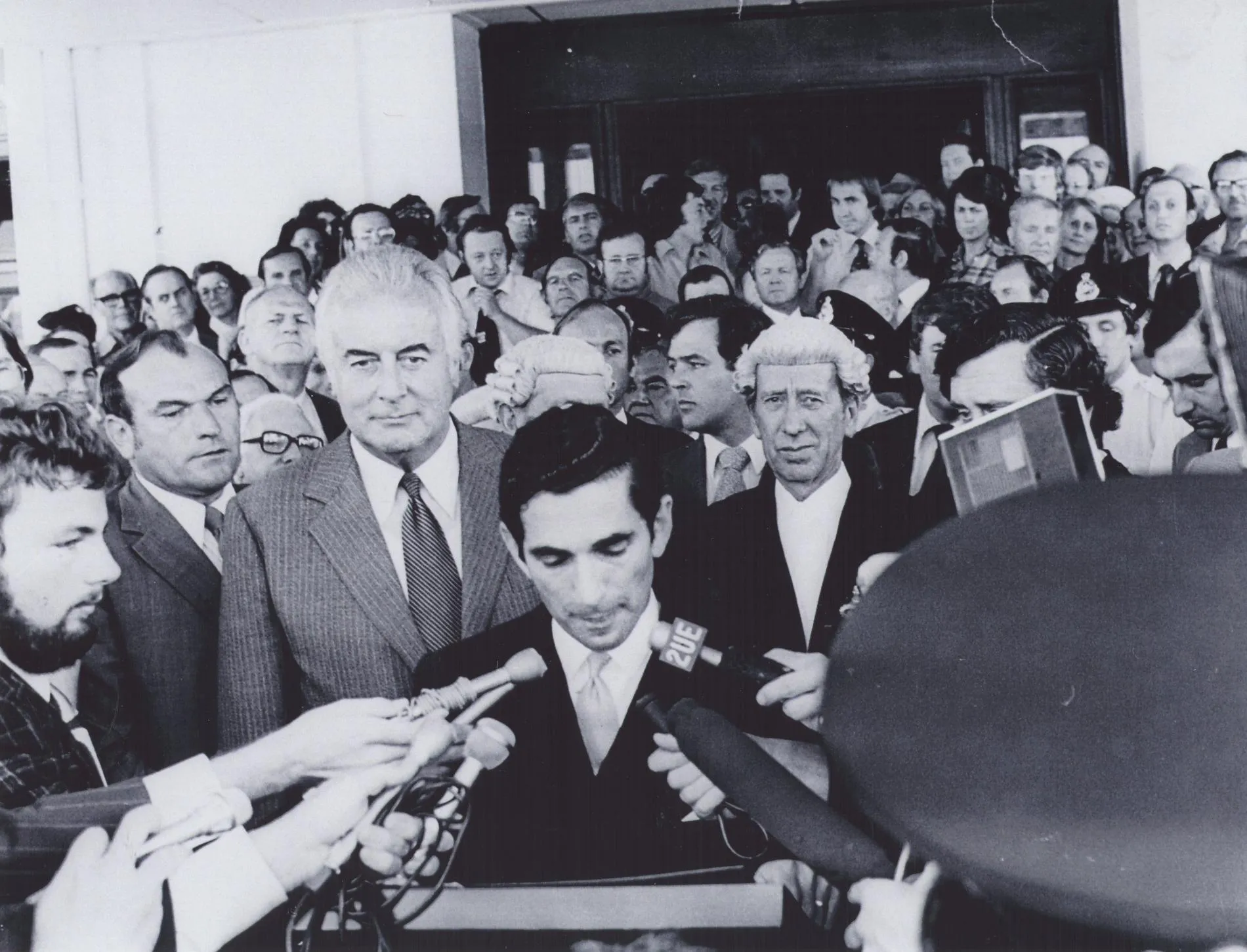David Smith reads the dismissal proclamation outside Parliament House in 1975. Gough Whitlam stands behind him amongst a huge crowd. Reporters hold microphones. David Smith reads the dismissal proclamation outside Parliament House in 1975. Gough Whitlam stands behind him amongst a huge crowd. Reporters hold microphones.